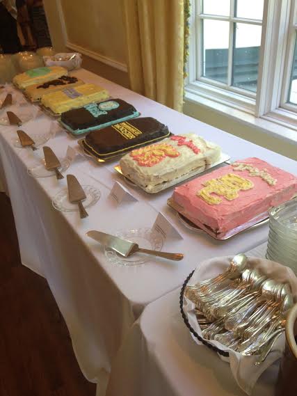 Seven graduation cakes decorated with college logos are displayed on a table